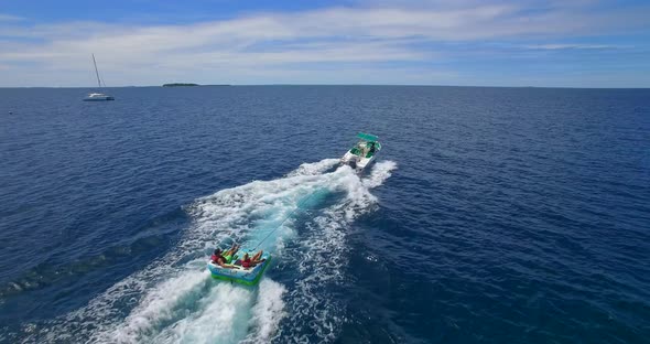 Aerial drone view of man and woman on an inflatable tube towing behind a boat to a tropical island alt
