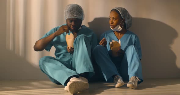 Afroamerican Doctors Eat Snack Together Sitting on Floor in Hospital Corridor alt