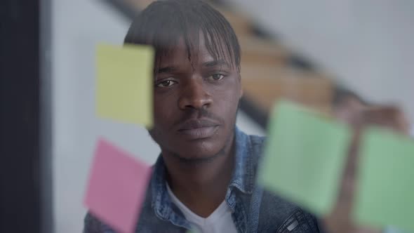 Handsome Young Man with Thoughtful Facial Expression Writing with Pen on Adhesive Notes on Glass alt