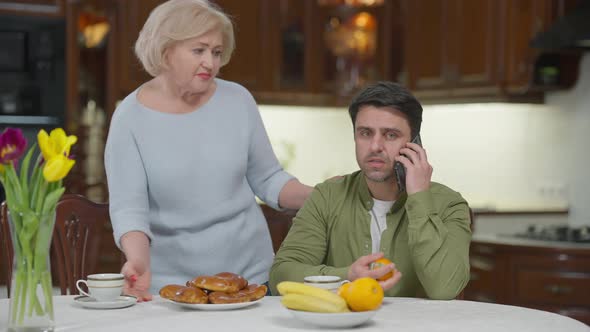 Busy Man Talking on Phone As Caring Senior Woman Bringing Dessert Shaking Head with Sad Facial alt