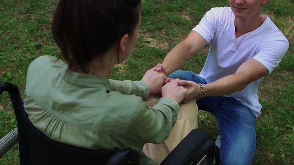 A Woman in a Wheelchair and Her Boyfriend on His Knees Holding Hands and Talking alt
