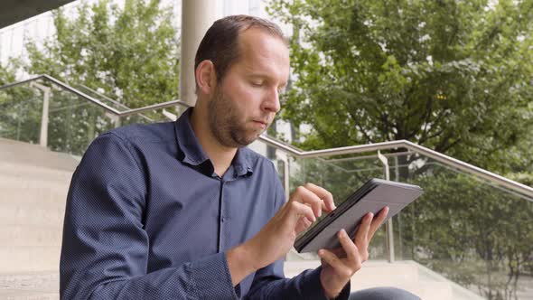 A Caucasian Man Works on a Tablet As He Sits on a Staircase in an Urban Area  Closeup From Below