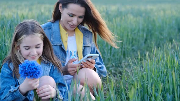 Mom and Daughter Use a Smartphone Sitting in the Tall Green Grass in the Middle of the Field alt