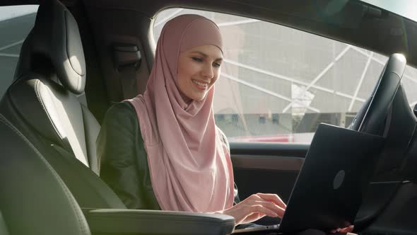 Attractive Business Lady in Hijab Sitting in Car with Laptop on Knees and alt