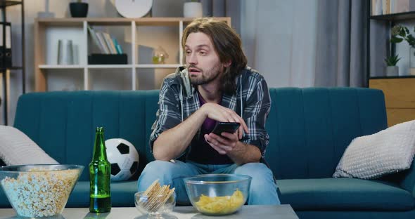 Guy is Sad while Watching Sport Game on TV at Home and Starting to Use His Mobile alt
