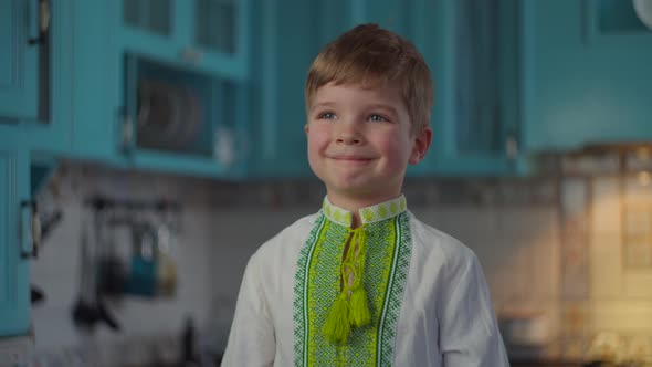 Blonde Boy in Embroidered National Clothes Smiling and Showing Emotions at Home on Blue Kitchen. Kid alt