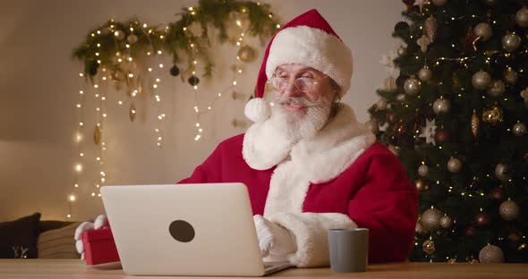 Elderly Bearded Man Dressed As Santa Sitting at Table with Laptop and Talking on Video Call Showing alt