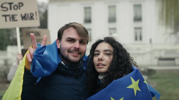 Embraced caucasian man and woman covered with EU and Ukraine flags are showing a peace sign. Shot wi alt