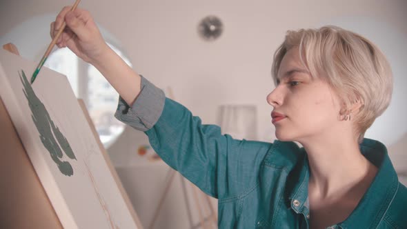 A Young Woman with Short Blonde Hair Painting with a Green Color in the Art Studio alt