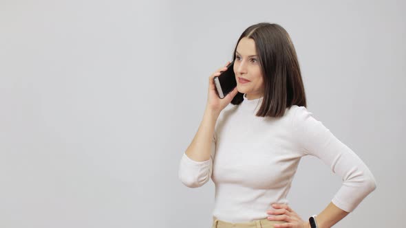 Joyful Woman In Beige Outfit Talking By A Cell Phone On A Grey Background, Copy Space