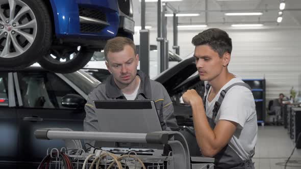Two Auto Mechanics Talking at the Garage Using Laptop in Car Diagnostics
