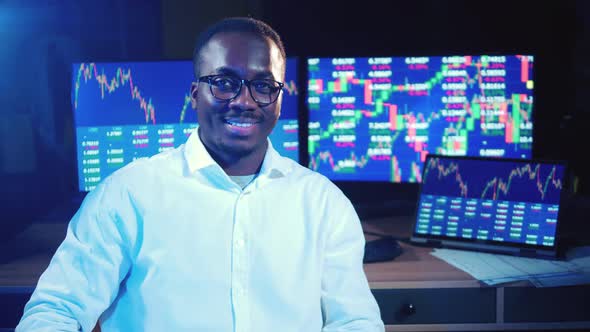 African Broker is Sitting Next to the Monitors with Stock Diagrams alt