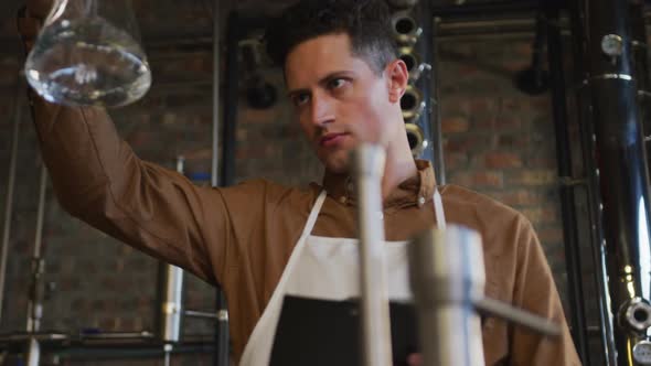 Caucasian man working at distillery checking gin product in flask and writing on clipboard alt