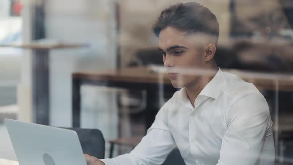 Young Businessman Using Laptop Computer at Cozy Coffee Shop. Window Reflection. He Is Sitting By the alt