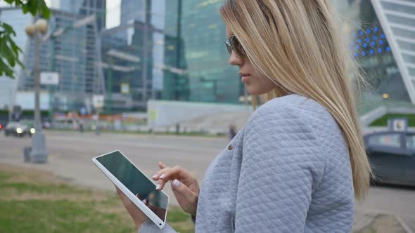 A Young Blond Girl Uses a Tablet on a Background of Skyscrapers Downtown alt