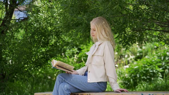 Carefree Young Woman Reading a Fascinating Novel While Sitting on a Park Bench alt
