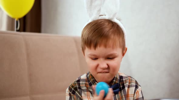 A Boy in Bunny Ears Holds a Blue Egg in His Hands and Depicts a Rabbit alt