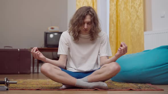 Portrait of Calm Young Retro Man with Long Curly Hair Falling Asleep Sitting in Lotus Pose Indoors alt