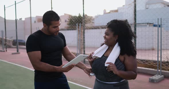 Curvy african woman talking with her personal trainer while checking tech clipboard outdoor alt