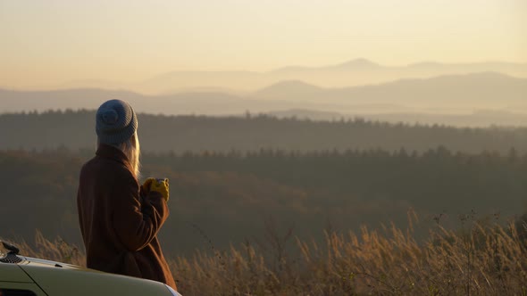 girl with mug of tea sitting on next to car in beautiful mountains Sudety in November in sunset
