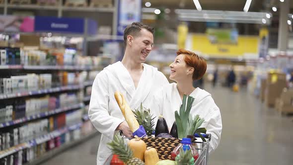 Young Couple in Bathrobes in Supermarket Doing Daily Shopping Walking with Cart alt