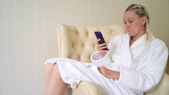 Young woman in white bathrobe uses mobile phone while sitting on armchair. alt