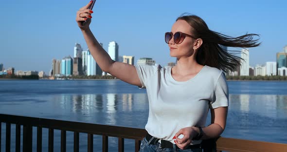 In Summer, the Girl on the Background of the Big City and the Waterfront Takes Selfie on Smartphone alt