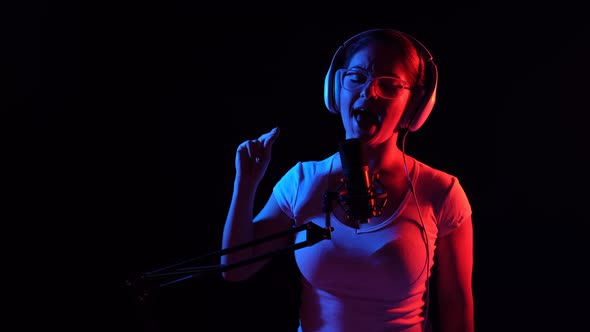 Caucasian Woman in Glasses and Headphones Sings Into a Microphone in Neon Light on a Black alt