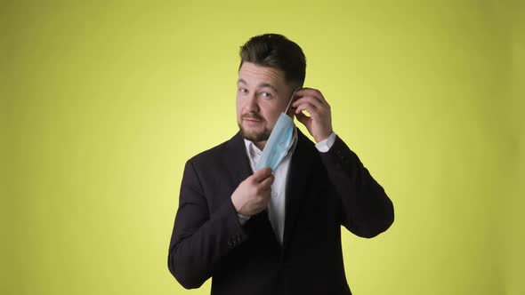 Young Bearded Man in Office Suit Putting on Mask on Face on Yellow Background