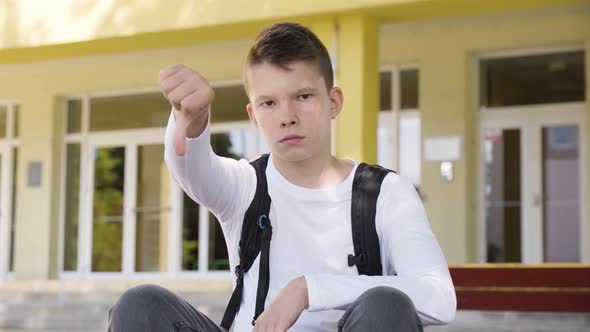 A Caucasian Boy Shows a Thumb Down to the Camera and Shakes His Head As He Sits in Front of School