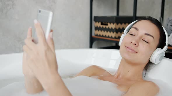 Joyful Student Wearing Headphones Listening To Music and Touching Smartphone Screen in Bathtub alt