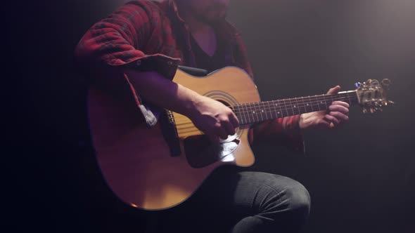 A male guitarist plays an acoustic guitar in a dark room.