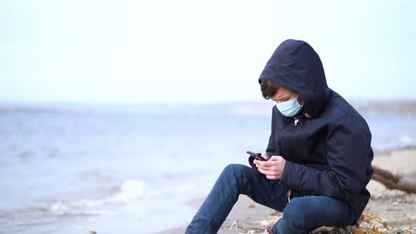 Teen Playing Online Video Games Sitting on Autumn Empty Beach During Pandemic Coronavirus Season alt