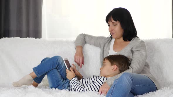 A Happy Mother and a Young Child Spend Time with a Tablet While Sitting on the Couch at Home. Mother alt
