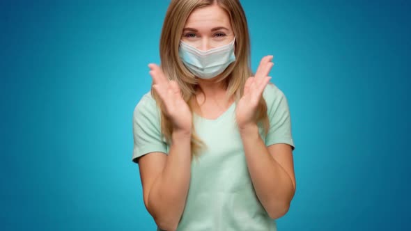 Woman in Medical Mask on Face is Very Happy and Claps Her Hands Against Blue Background alt