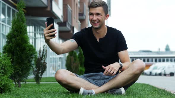 Handsome Young Man Talking in Video Chat Via Cell Phone, Sitting on the Grass and Speak with Friends alt