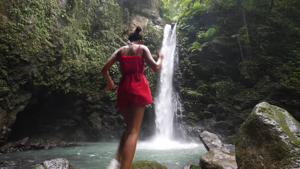 Young Woman in a Red Romper Walks and Jumps at the Foot of a Waterfall alt
