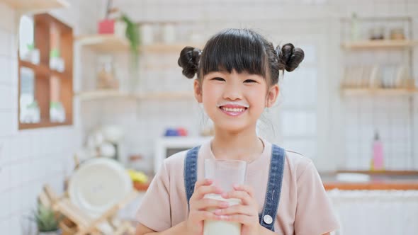 Portrait of Asian little cute preschool kid holding a cup of milk and drinking in kitchen at home. alt