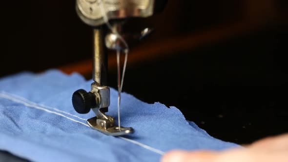 The Seamstress Sews With A White Thread On An Old Sewing Machine A Blue Cloth On A Dark Background