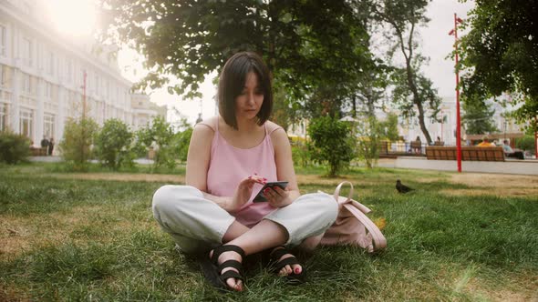 Young Asian woman sitting on grass in the park holding smartphone, using mobile apps. Stylish girl. alt