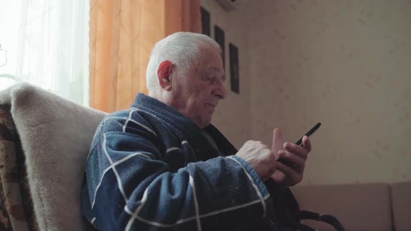 Elderly Man with Smartphone Sitting in an Armchair and Scrolling Social Media alt