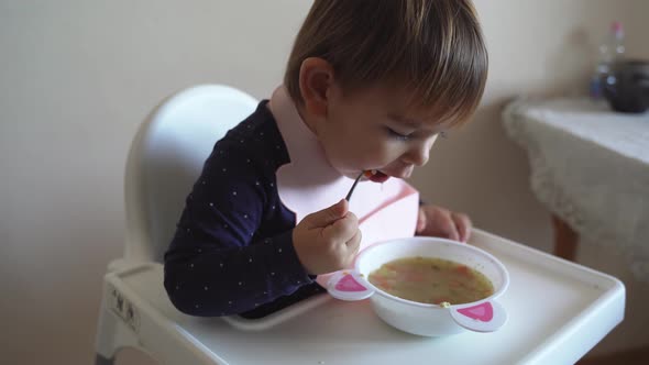 Young Toddler Girl Witth Silicone Bib Eating Alone Soup with Spoon alt