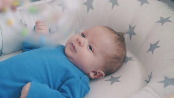 Little Child Looks at Rattle in Mom Hand in Sleeping Cocoon alt