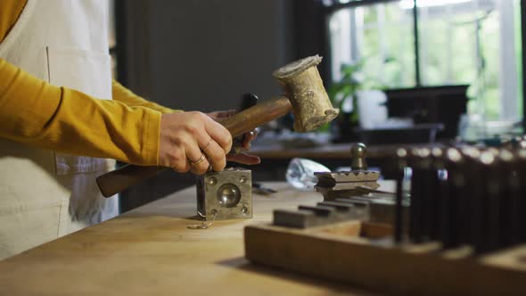 Midsection of caucasian female jeweller in workshop wearing apron, using hammer, making jewelry alt