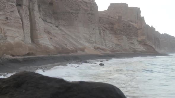 Tracking shot of waves hitting a black beach that's surrounded by tall white volcanic cliff formatio alt
