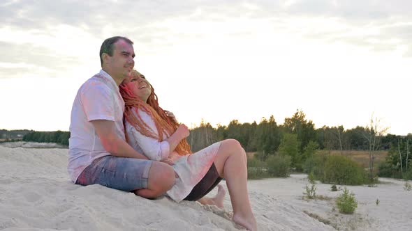 Cheerful Young Couple Sitting on the White Sand. Faces and Clothes Are Painted in Bright Colors alt