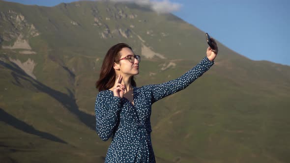 A Young Woman in a Dress Stands in the Mountains and Takes a Selfie on a Smartphone. alt