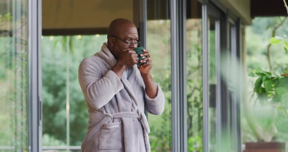 African american senior man standing on balcony wearing bathrobe drinking coffee and enjoying view alt