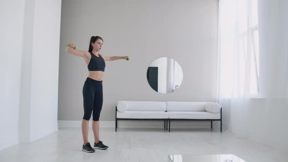 A Woman Raises Her Arms with Dumbbells Doing a Shoulder Exercise in Her Light White Apartment alt