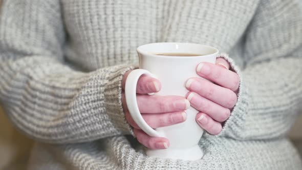For lack of heating, woman wearing a warm sweater warms hands, holding mug with hot tea. Close up